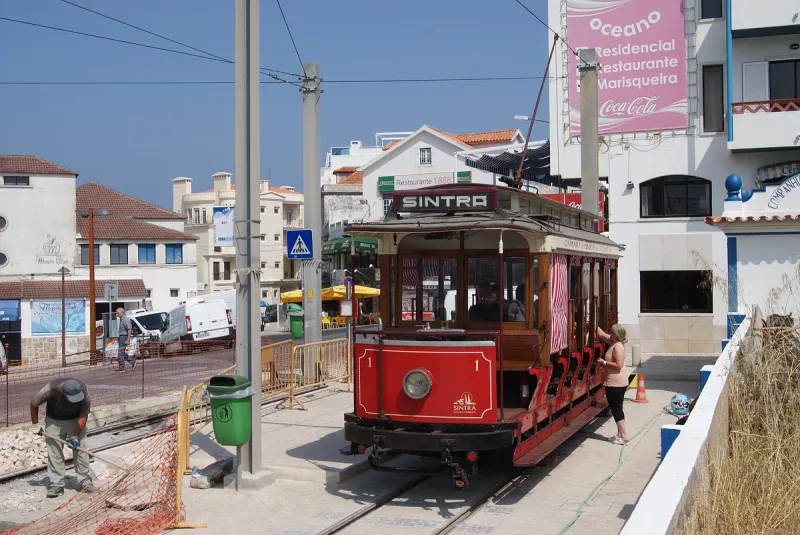 חשמלית עץ אדומה ועתיקה עומדת על פסים ברחוב, ובניינים לבנים משני צדיה