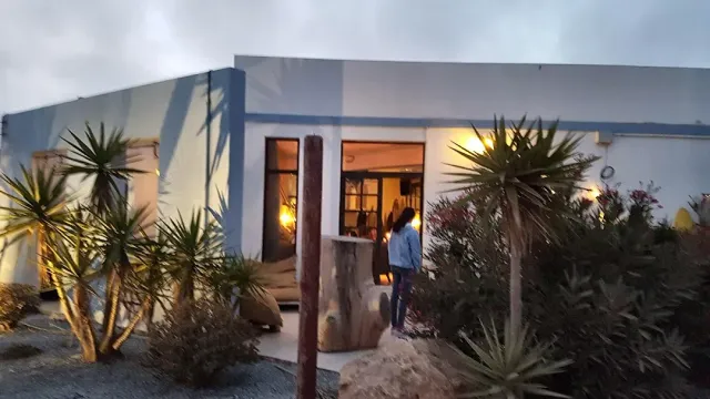 מבנה נמוך בלבן וכחול בשעת דמדומים, פתח מואר וצמחי יוקה בחזית