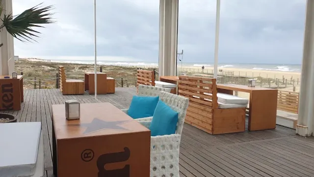 מרפסת עץ עם ספות וכריות טורקיז מול חוף חולי וים אפור