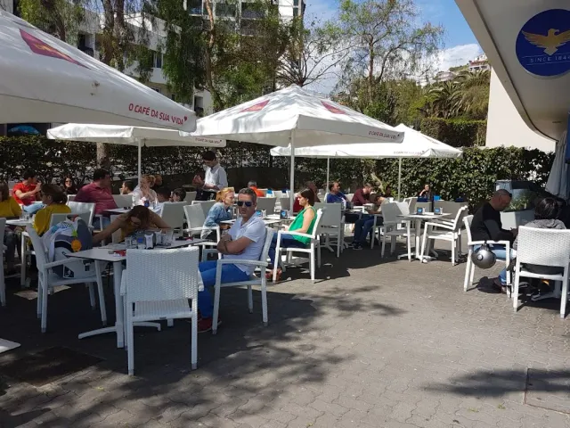 מרפסת עמוסה סועדים תחת שמשיות לבנות, כיסאות פלסטיק לבנים ועצים ברקע
