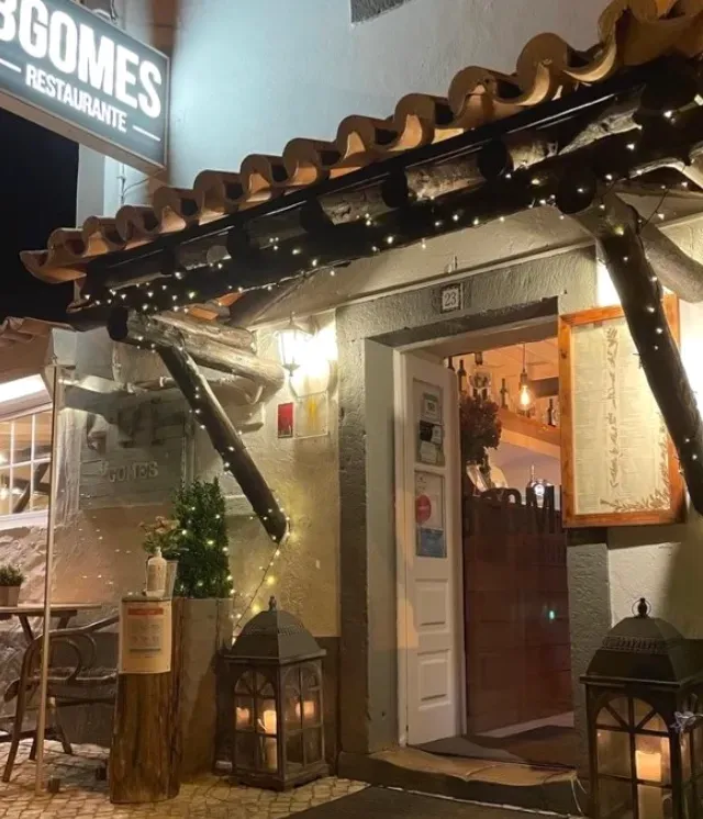 כניסה בלילה עם סככת עץ, שרשרת נורות קטנות ופנסים דולקים ברצפה