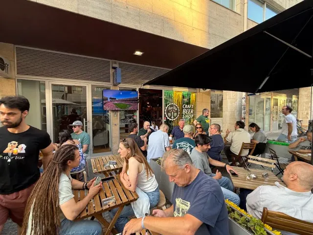 רחבה עמוסה באנשים יושבים על ספסלי עץ מול חנות בירה