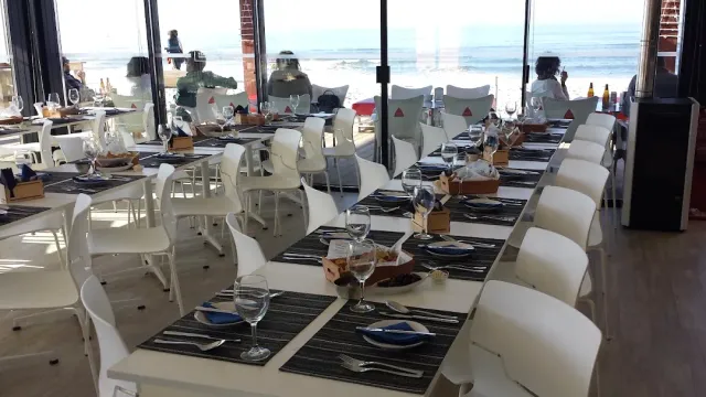 שולחנות ארוכים עם כיסאות לבנים מול קיר זכוכית אל החוף והים