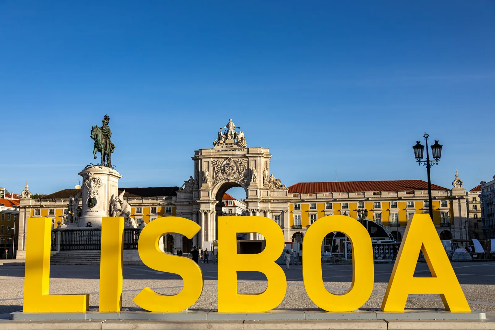 אותיות LISBOA צהובות וגדולות בכיכר קומרסיו בליסבון, על רקע שער הניצחון ופסל המלך ז'וזה הראשון, באור יום